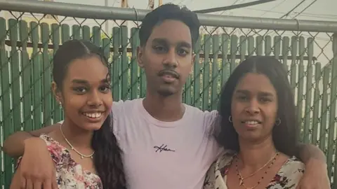 Family Shawn Seesahai with his arms around his sister and mother; They all smile at the camera