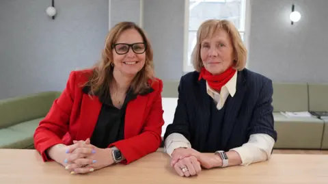 Helen Godwin and Catherine Gibbons sit next to each other at a table, smiling at the camera. Helen is wearing a red suit jacket and Caherine is wearing a red neck scarf.