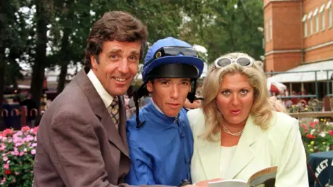 PA Media  John Stapleton, wearing a brown suit, smiles alongside jockey Frankie Dettori, wearing blue silks, and Vanessa Feltz at Newmarket race course