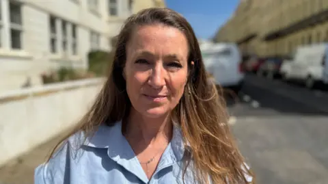 Marianne stands with a view of Oriental Place, Brighton, behind her. She has long light brown hair and wears a light blue shirt with a silver necklace.