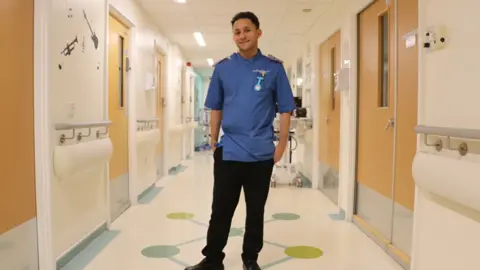 A man in a nurse's uniform stands in a hospital corridor.