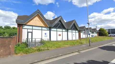 A building with several pointed gables is boarded up with overgrown grass in front of it next to a pavement and road. The sun is shining.
