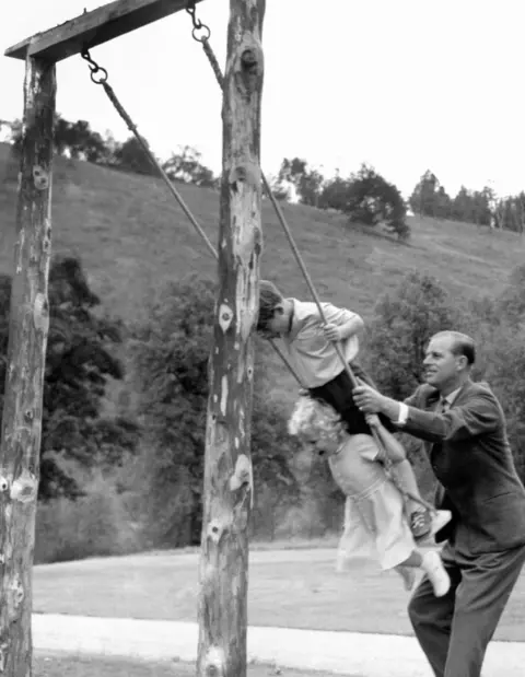 Buckingham Palace A black and white image of Prince Phillip pulling back the ropes of a swing upon which a young King Charles and Princess Anne are standing.