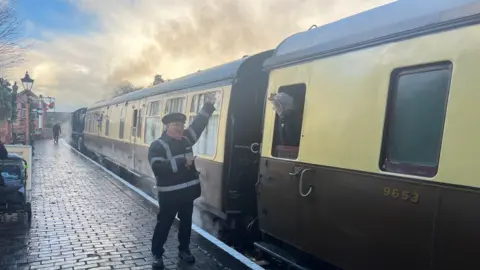 A guard wearing a black and white uniform on the platform is holding his left arm up standing on a platform by a yellow and brown train.