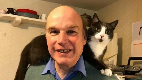 Charles Morris Charles Morris is smiling and wearing a blue shirt and green jumper. He has a black and white cat on his back. 