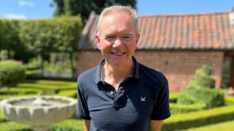 Shaun Whitmore/BBC Dr Paul Everden a man who is standing outside in his garden. He is standing in front of leafy green bushes and hedges. He is looking directly at the camera and smiling and is wearing a navy polo shirt.
