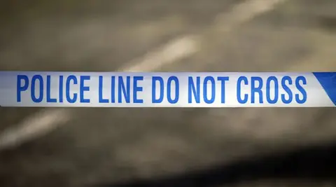 PA Media Generic picture of a blue and white police tape stretched across a road with the words Police Line Do Not Cross
