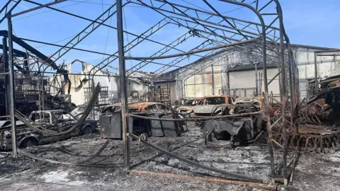 The burnt-out frames of warehouses and cars stand on a site covered in charred wreckage from the motorsport business. The frames are warped from the heat of the fire and the whole site is covered in ash.