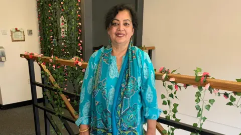 A South Asian woman stands at the top of a staircase which is decorated with trailing green leaves and flowers. She is wearing a dark green tunic with a lighter green pattern all over and a darker green scarf draped around her shoulders. she has dark hair pulled back from her face and is smiling into the camera.
