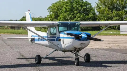 Getty Images A small plane that is white with blue stripes and has a propeller on the front. It is on a runway in front of trees on a sunny day.