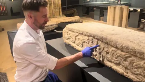 BBC/ Emily Johnson A man in a white shirt and blue gloves points to carvings in stonework.