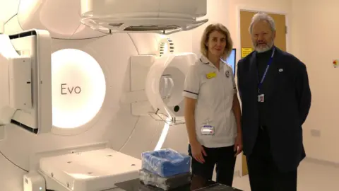 A man and female clinician standing next to a radiotherapy machine in a hospital environment. The walls and machine are white. The man has white hair and beard and is wearing a black suit, black polo neck and lanyard. The female is in a white medical jacket with short sleeves and blonde shoulder-length hair.