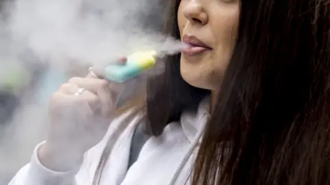 EPA A close up of a young woman using a disposable vape. Only the bottom half of her face can be seen. She is wearing a white hoody. 