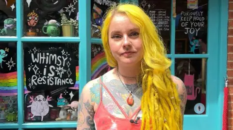 Image of Katie. She has yellow hair and lots of colourful tattoos.