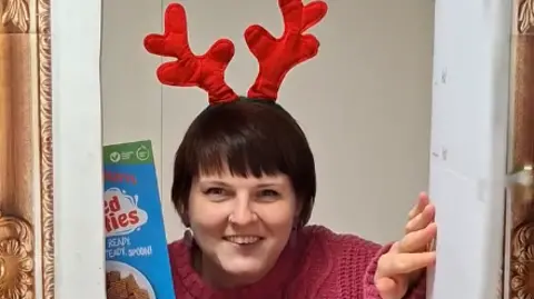 Redcar Area Foodbank A woman with short, dark hair, dangly earrings and a pink jumper standing behind the same frame. She is holding a blue box of cereal in one hand and her other is holding onto the white door of the frame.