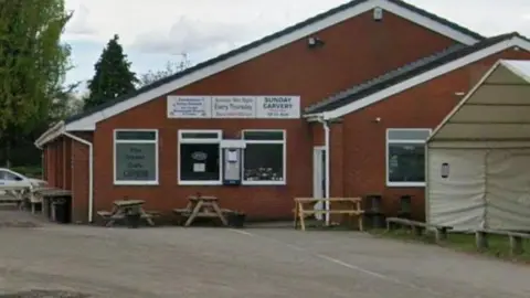 A small brown building with a slanted roof. There are several wooden tables outside and part of a white marquee can be seen to the right. 