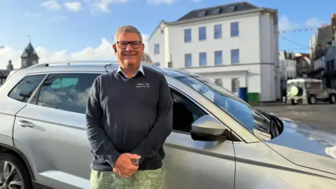 BBC Photo of a man standing in front of a silver taxi. He is wearing a navy jumper with a blue collar poking out the top. He has short grey hair and has black glasses on