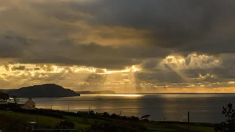 MANX SCENES Sunset at Niarbyl Bay on a cloudy day. The remaining yellow light is streaming through the clouds illuminating part of the water below. A silhouette of the Calf of Man in the distance.