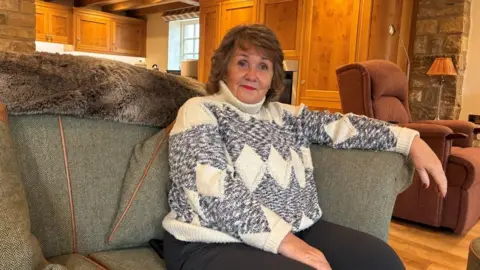 The image shows a woman seated on a large, cushioned sofa.
She is wearing a patterned knitted sweater with geometric shapes in light and dark shades, along with dark trousers.