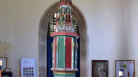 Simon Knott The font is in the centre of the image with an archway behind. It is very ornate and coloured in red, green and gold