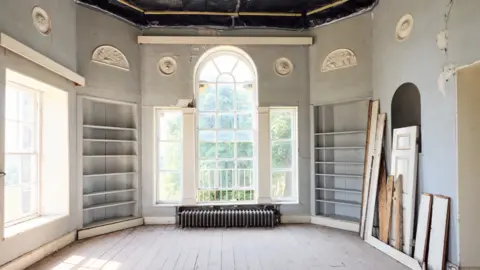 Olivia Brabbs/Landmark Trust A sun filled room painted in pale grey. The room is dominated by a large central window made up of three large panes, two thin on either side of a larger central pane through which the light is pouring in. Outside greenery is visible. Underneath the window is a long, grey rectangular radiator which rests on wooden floorboards. On either side of the window are empty built in shelves. Above the shelves just below ceiling level are 6 white Wedgwood style friezes. On the right hand side are pieces of wood and an unhinged cupboard door.