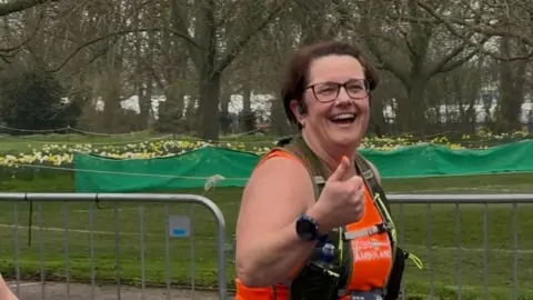 SAMANTHA CLARKE Samantha Clarke is smiling and offering a thumbs up to the camera as she runs the Cambridge Half Marathon. She is wearing an orange T-shirt and running vest holding a water bottle. Behind her is metal railing and behind that is grass, daffodils and trees. 