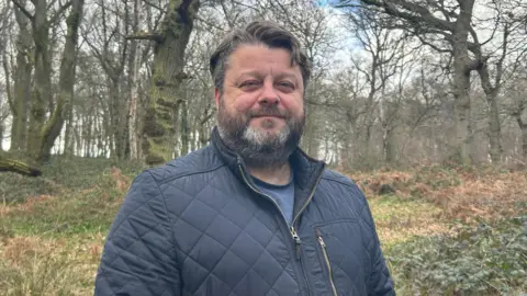 A man standing outdoors in a woodland setting. The man is wearing a dark quilted jacket with a zip and a high collar, along with a darker top underneath. The individual is positioned slightly off‑centre, with one arm resting by their side.