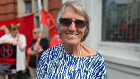 Elaine Murray has short grey hair, is wearing sunglasses, and a blue, white and green patterned blouse. She is standing in front of white and brick buildings, with protestors behind to the left with placards and flags.