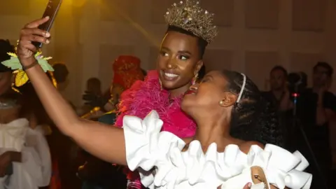  Getty Images Two women at the "Bridgerton Affair" party smiling for a selfie. One of the women in wearing a crown and an elaborate pink dress. The other woman is dresses in a white outfit.