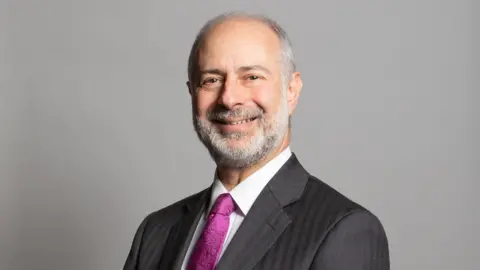 UK Parliament Leeds North East MP, Fabian Hamilton has grey hair and a beard. He wears a suit with a purple tie.