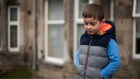 Getty Images A sad-looking child with dark hair, wearing a grey body warmer over a blue top, looks at the ground while standing in a street 