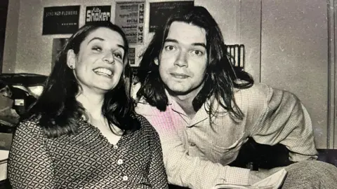 A black and white photo of Conal with his wife Lee. Conal has long dark hair and is wearing a shirt and his wife has long brown hair and a patterned button up blouse. They are both looking at the camera.