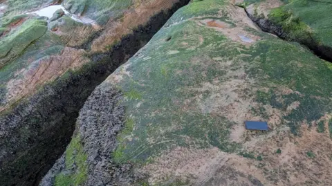 Wirral Coastguard A narrow crevice runs between large rocks coated in green algae and brown seaweed. A black smartphone is visible on the surface of the rocks in the foreground. 
