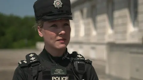 Róisín Brown wearing a dark green police uniform standing outside a light stone building. She is wearing a police cap and her hair is tied back. 