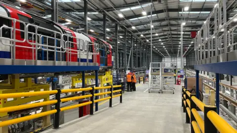 Pritti Mistry/BBC A view inside the factory. A ramp leading down to the main floor has yellow rails either side. To the left is a red, white and blue tube carriage propped up on a stand and three workers wearing orange hi-vis vests are talking in the background.