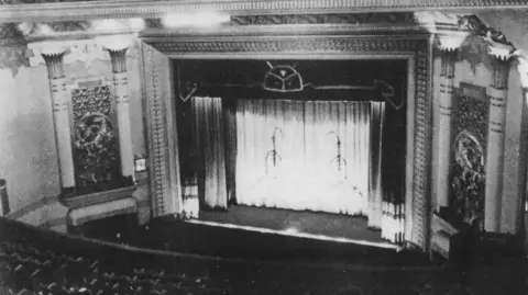 FCSA An old, black and white photo of the Forum auditorium which has a circle and stalls below. Either side of the curtained stage are twin pillars. 