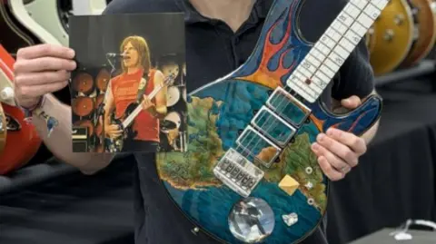 Gardiner Houlgate/PA A close-up image of a person's hands holding a rare blue guitar with orange flames on its body and images of the earth, with a thermometer up its stem next to a photograph of a singer holding the guitar. 