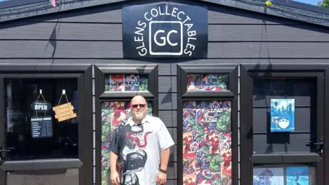 Supplied A man wearing a Venom shirt standing in front of his shop, which is a black building with comic book characters plastered over the front windows. 