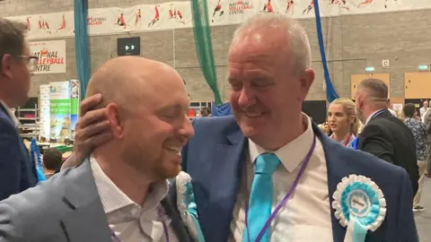 Martin Griffiths smiles and holds the back of a fellow councillor's head. He is wearing a light blue tie, white shirt and navy suit jacket while smiling. He is standing inside a sports hall where local election votes are being counted.