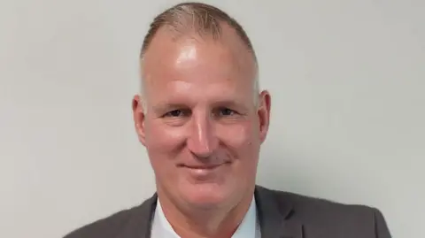 Shropshire Council A short-haired man wearing a jacket and white shirt is smiling in front of a plain, grey background.
