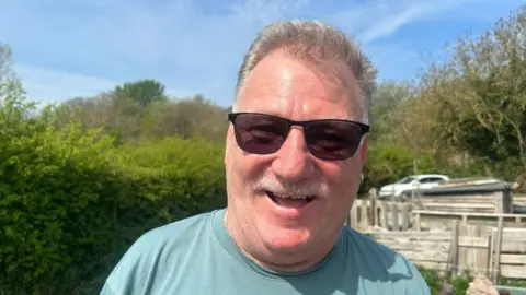 A man wearing sunglasses, a green T-shirt and an earring smiles at the camera.