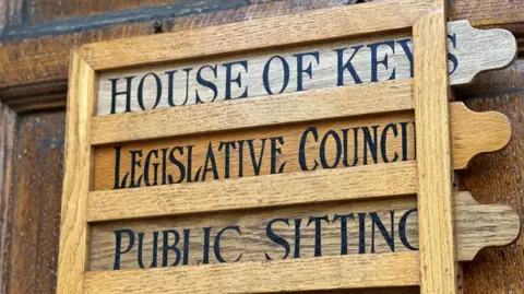 A wooden sign on the door of the parliament building that reads House of Keys, Legislative Council and Public Sitting.