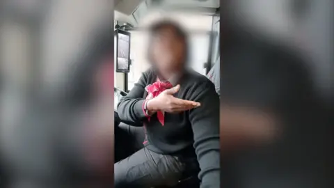A bus driver gestures with one hand while wearing a black sweater and a red bow tie.