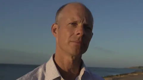 Local Democracy Reporting Service A bald man in a shirt with squares on it. There is a beach and the sea in the background.