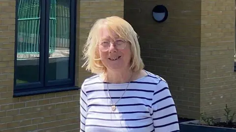 Julie Young, who has short blonde hair, wears a black and white-striped top in front of a cream brick building. She is smiling and wearing glasses.