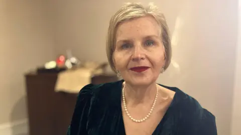 The head and shoulders of a woman with blonde bobbed hair, wearing red lipstick, pearl and silver earrings, a pearl necklace and navy velvet top. She is seated in a room with beige walls and a brown dresser out of focus behind.