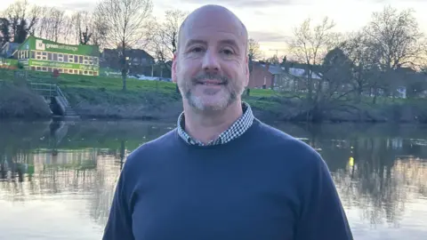 Gavin Kermack A bald man with a short salt-and-pepper beard stands in front of a river, smiling at the camera. He is wearing a blue-and-white check shirt underneath a dark blue sweater.