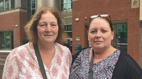 Two women in their 50s stand next to each other outside a court building. It is a close up shot. They have neutral or slightly smiling expressions. Linda on the right is slightly older, with a patterned white and pink blouse and short curly strawberry blond hair. Deborah on the right is shorter with a patterned blue and white top, black cardigan, and brown hair in a ponytail.