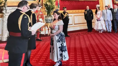 An investiture ceremony taking place at St James's Palace. Ann Marie-Riley, wearing a dress and hat, can, is standing in front of Princess Anne while others look on in the background. The room has a large red carpet and contains many regal decorations.