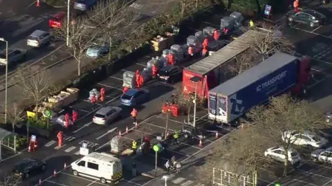 Aerial look at car park with queue of cars and people in red outfites lined up by pallets of water.